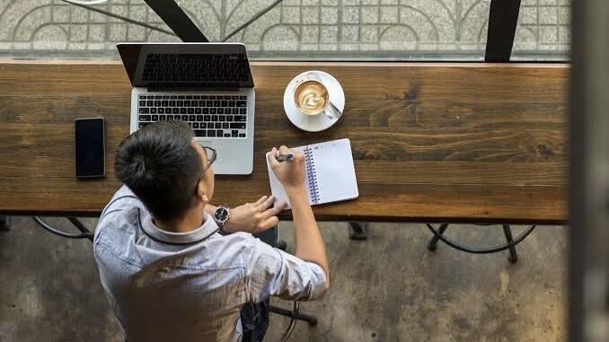 Man sitting at a table with laptop and coffee, writing in sketchpag